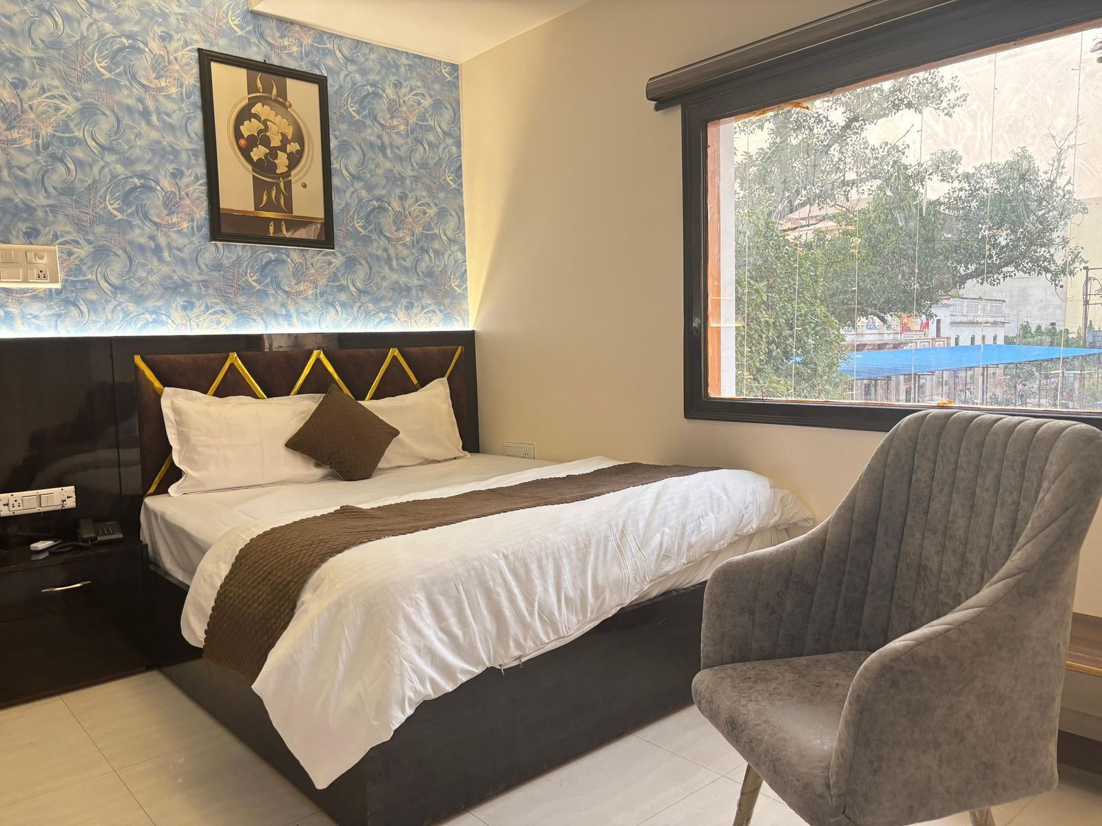 Quarto de hotel com cama, cadeira, janela e vista para edifícios, além de uma imagem emoldurada na parede.