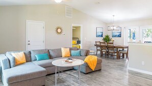 A modern living room with a sectional sofa, a dining table, and a ceiling fan. - Family Beach Retreat w/ Private Fenced Backyard, Fire Pit, Games (Englewood)