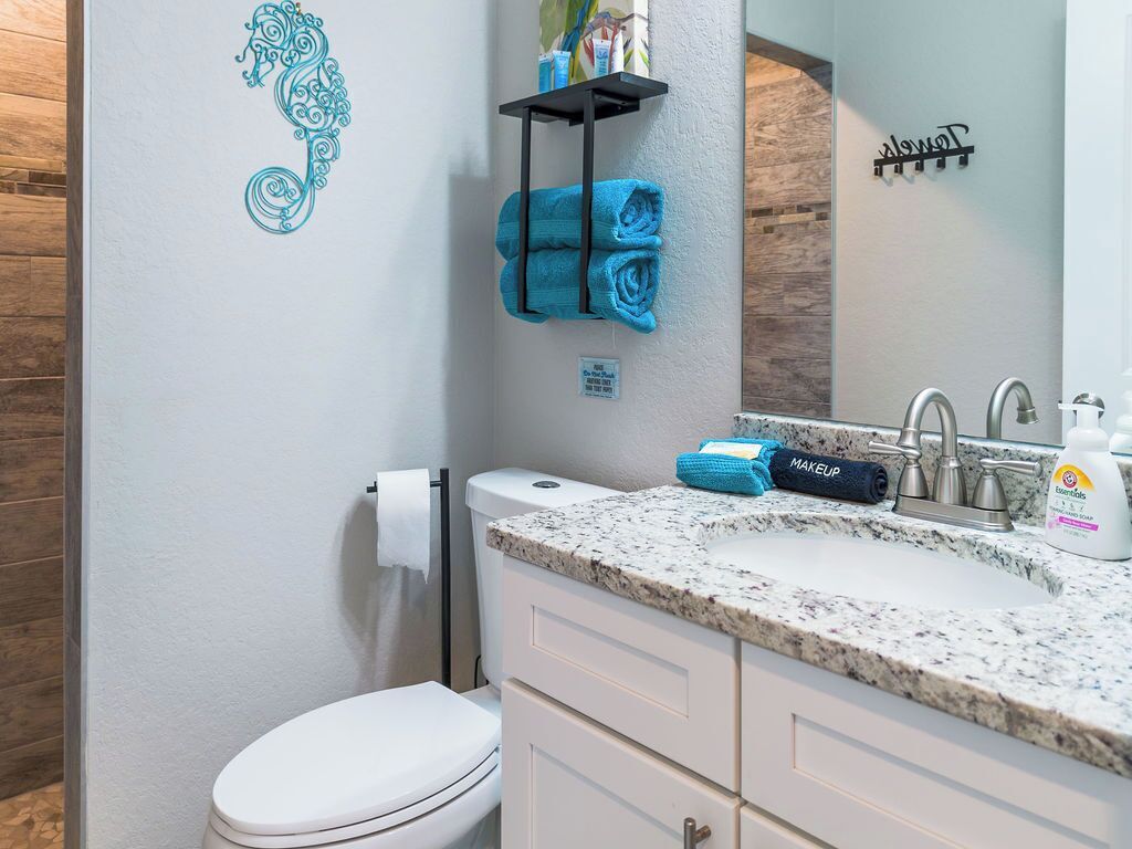 A bathroom with a toilet, a sink with a granite countertop, a towel rack with blue towels, and a mirror with a towel rack above it.
