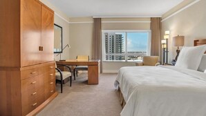 A hotel room with a large bed, a desk with a chair, a wardrobe, and a window with a city view.