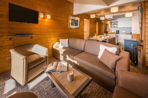 A cozy living room with a sofa, armchair, and coffee table, a flat-screen TV mounted on the wall, and a kitchen area visible through an open doorway. - Woodsy Retreat in the Alps (Albiez-Montrond)