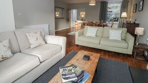 Ein Wohnzimmer mit einer Couch, einem Couchtisch mit Büchern, einem Essbereich mit Tisch und Stühlen sowie Blick ins Freie durch ein Fenster.