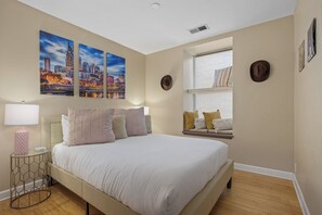 Une chambre à coucher comprenant un lit, une table de chevet, une fenêtre avec des stores et trois tableaux représentant des paysages urbains accrochés au mur.