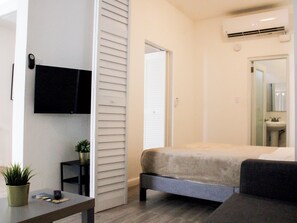 Een slaapkamer met een bed, een aan de muur bevestigde televisie, een nachtkastje met een plant, en een badkamer met een wastafel en spiegel.