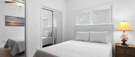 Un dormitorio con cama, mesita de noche, lámpara, ventana con persianas, espejo y un cuadro en la pared.