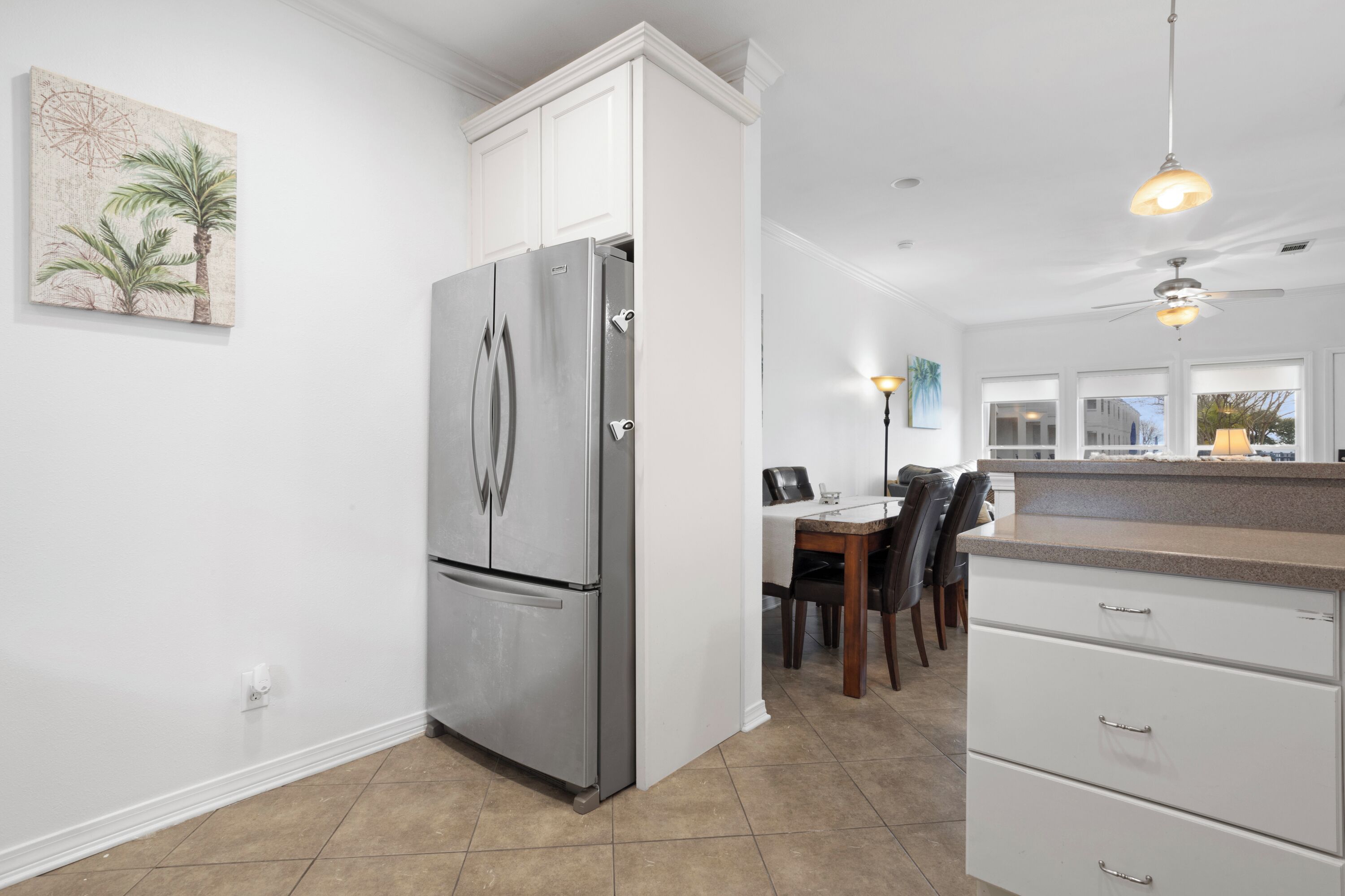 A modern kitchen with white cabinetry, a stainless steel refrigerator, a dining area with a table and chairs, and a view of the outside through large windows.