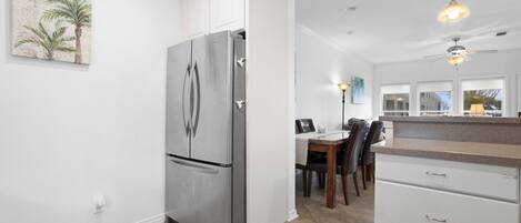 A modern kitchen with white cabinetry, a stainless steel refrigerator, a dining area with a table and chairs, and a view of the outside through large windows.