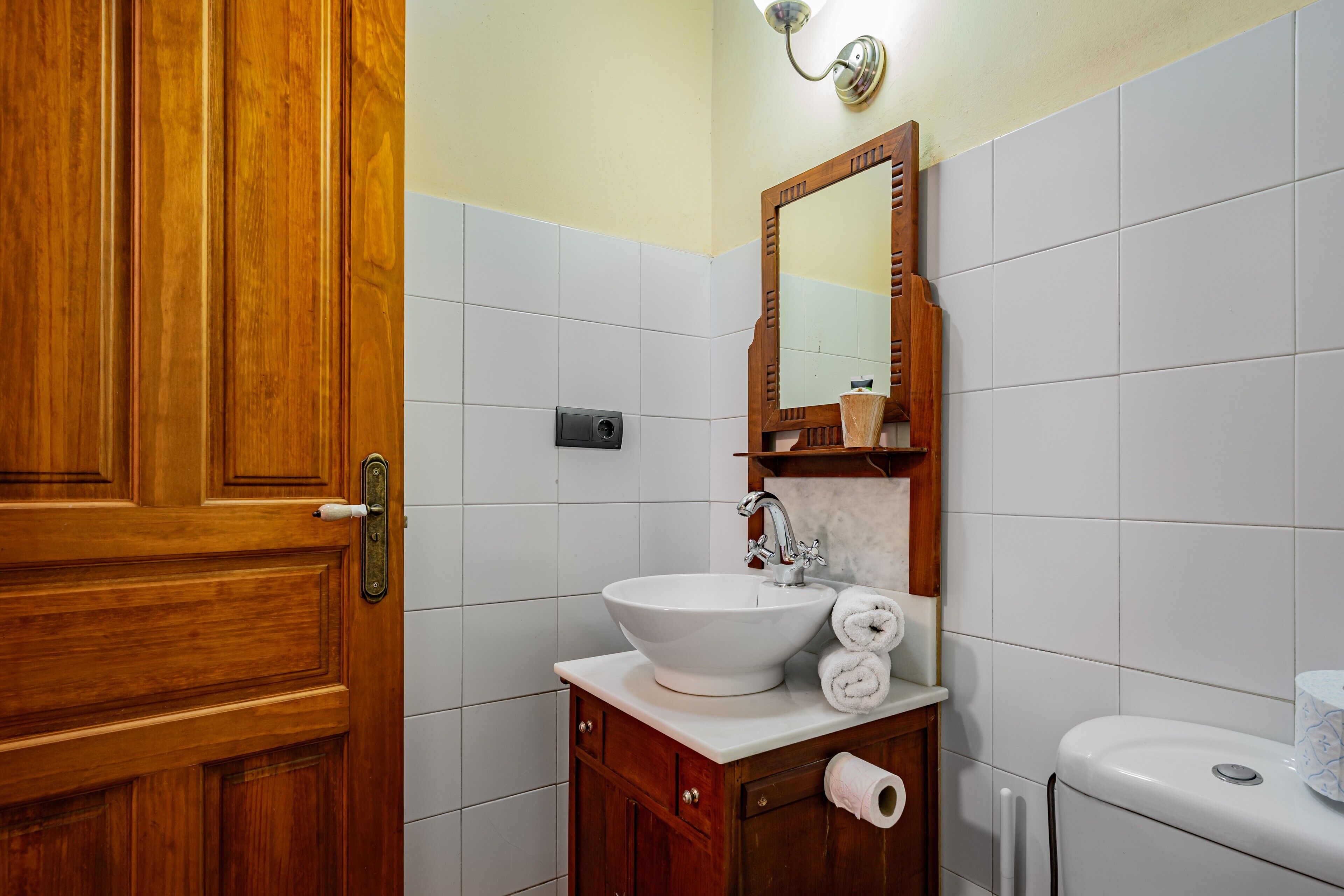 Un baño con puerta de madera, azulejos blancos, un lavamanos con espejo y un inodoro.