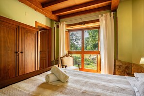 Une chambre avec une grande armoire en bois, un lit avec du linge de maison blanc, une chaise et une fenêtre donnant sur un paysage verdoyant.