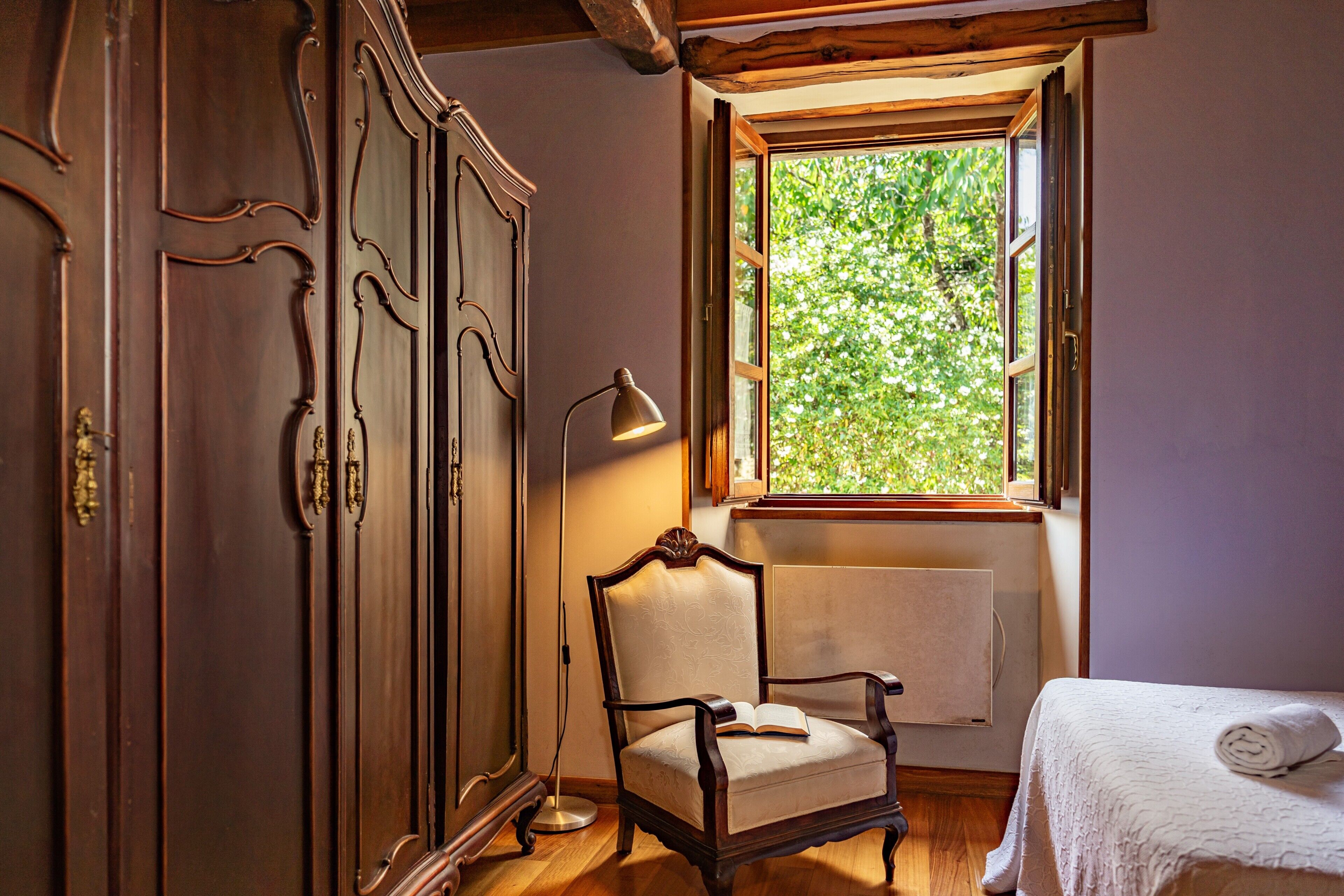 Habitación con armario de madera, una silla, una mesita de noche, una lámpara y una ventana con vistas a zonas verdes.