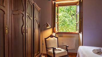 Habitación con armario de madera, una silla, una mesita de noche, una lámpara y una ventana con vistas a zonas verdes.