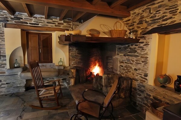 A stone fireplace with a fire, wooden chairs, a wooden door, and a stone wall.