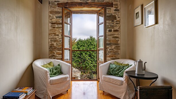 Una habitación con dos sillas blancas, una mesa con tetera, una estantería y vistas a través de una ventana enmarcadas en piedra.