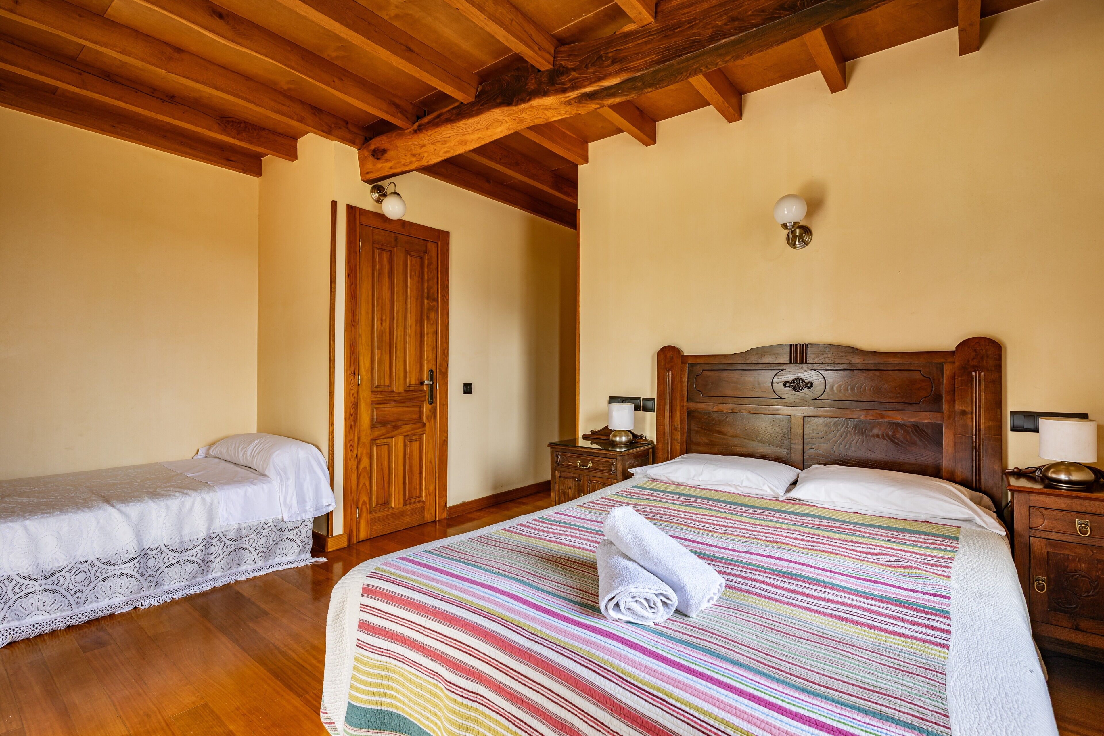 Un dormitorio con techo de madera, una cama grande con colcha a rayas de colores, una cama más pequeña, mesitas de noche de madera y una puerta de madera.