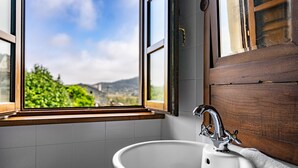 Un baño con un lavamanos blanco, una puerta de madera y una ventana con vistas a la vegetación y montañas.