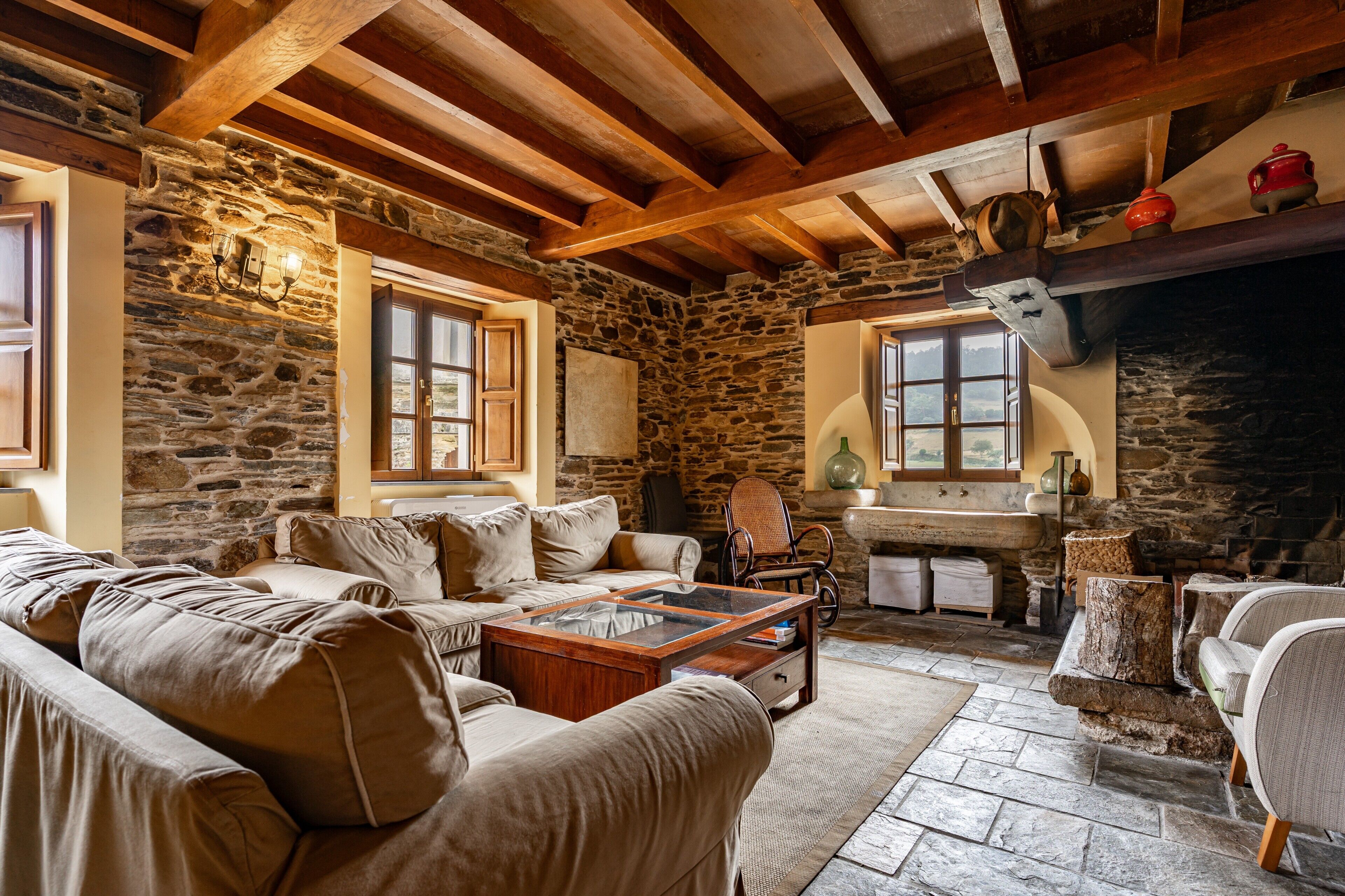 Un intérieur rustique avec des murs en pierre, des poutres en bois, une cheminée en pierre et un coin salon confortable avec un canapé et des fauteuils.
