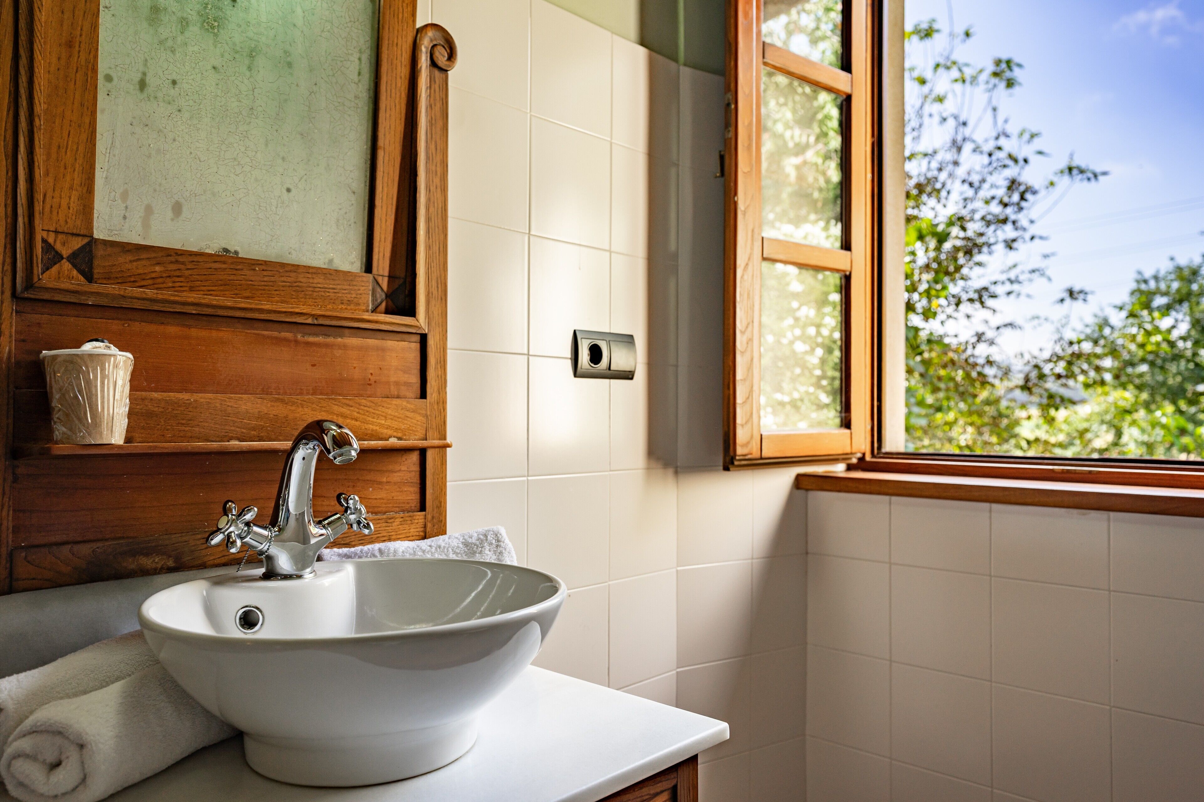 Un baño con lavamanos blanco, armarios de madera, una ventana y vistas al verdor exterior.