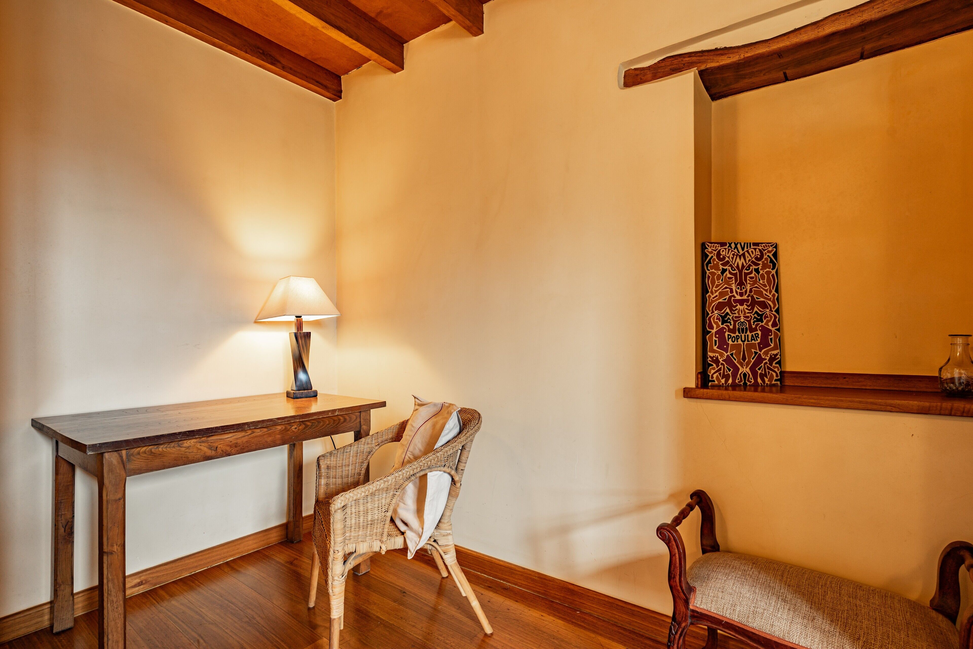 Una habitación con una mesa de madera, dos sillas de mimbre, una lámpara y un estante en la pared con un adorno.