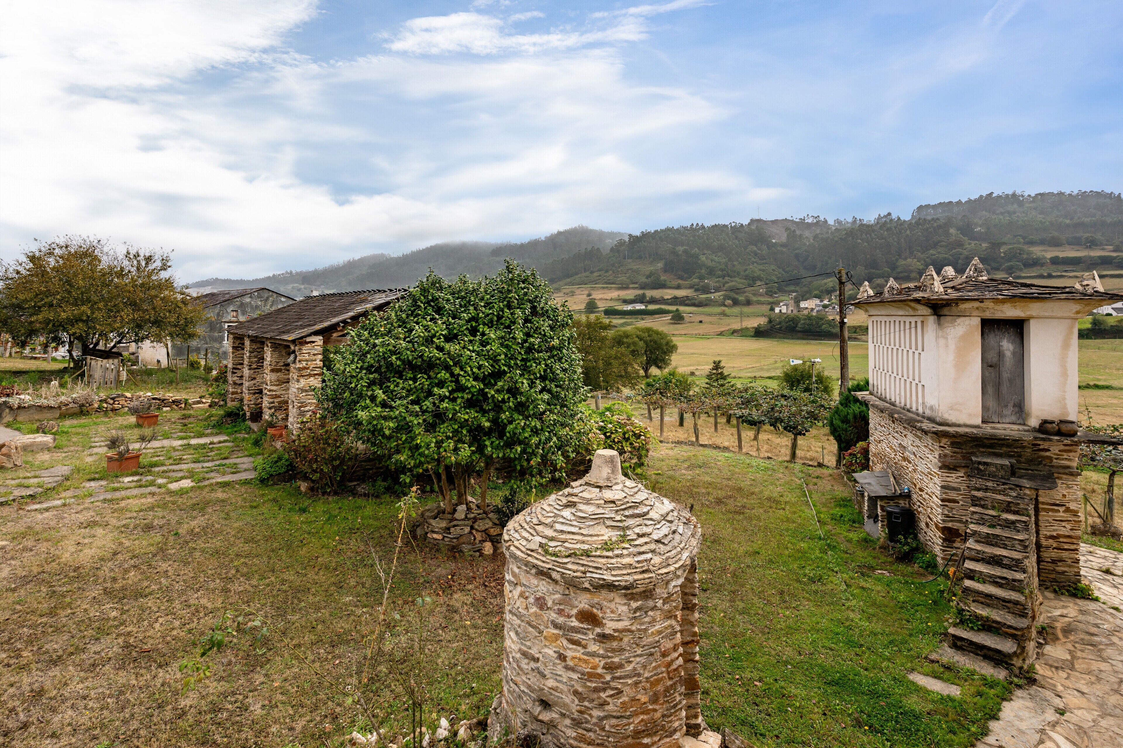 Un edificio rústico de piedra con techo de paja, rodeado de un área con césped, árboles y una cadena montañosa distante.