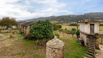 Un edificio rústico de piedra con techo de paja, rodeado de un área con césped, árboles y una cadena montañosa distante.