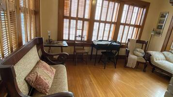 A spacious room with a wooden floor, a sofa, a dining table with chairs, and large windows with blinds.