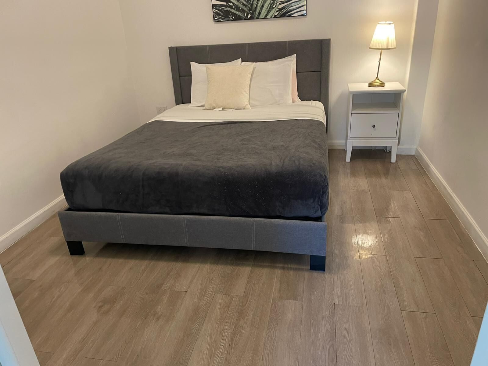 A neatly made bed with a grey headboard and white bedding, a bedside table with a lamp, and a wooden floor.