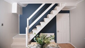 Una escalera con peldaños blancos que conduce a una puerta, un jarrón con plantas sobre una mesa y una lámpara colgante.