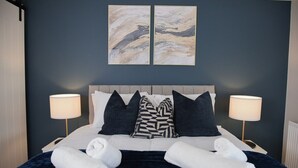 A neatly made bed with white linens and two towels, a dark blue headboard, and two bedside lamps. - Stylish 3-Bed • Hot Tub • Free Parking • Contractors (Essex)