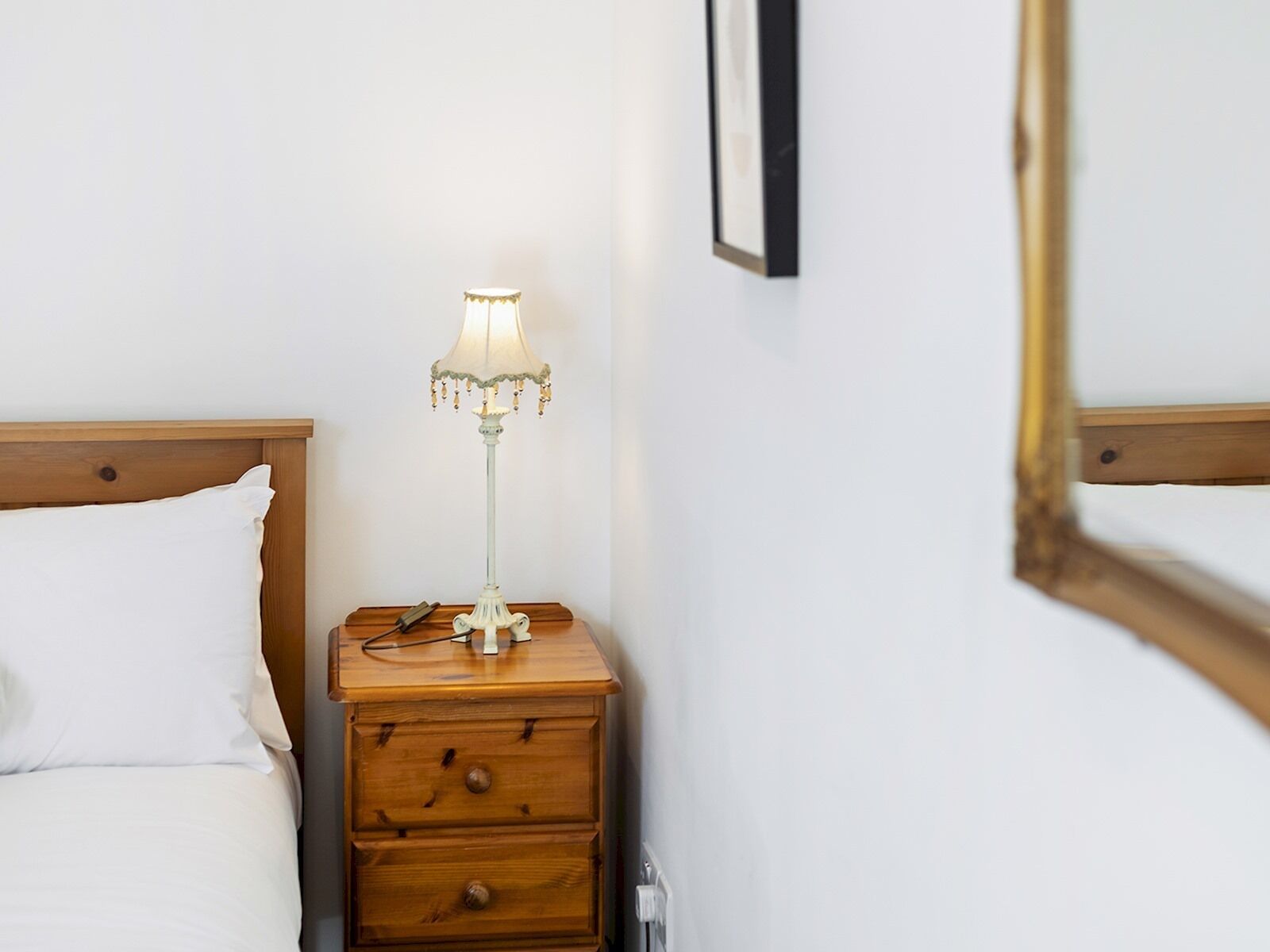 Een net opgemaakt bed met witte lakens, een houten nachtkastje met een lamp en een lijst met een afbeelding aan de muur.