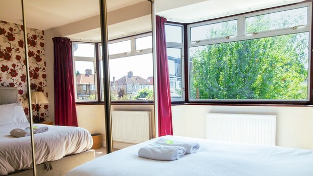 A hotel room with two beds, a large window with a view of greenery and buildings, and a mirror reflecting part of the room. - Cosy Home w/ Stylish Interior – PassTheKeys (Watford)