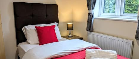 Una cama bien hecha con ropa de cama blanca y roja, una lámpara de noche y una ventana con cortinas.