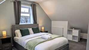 Quarto com cama, mesas de cabeceira, radiador e janela com cortinas.