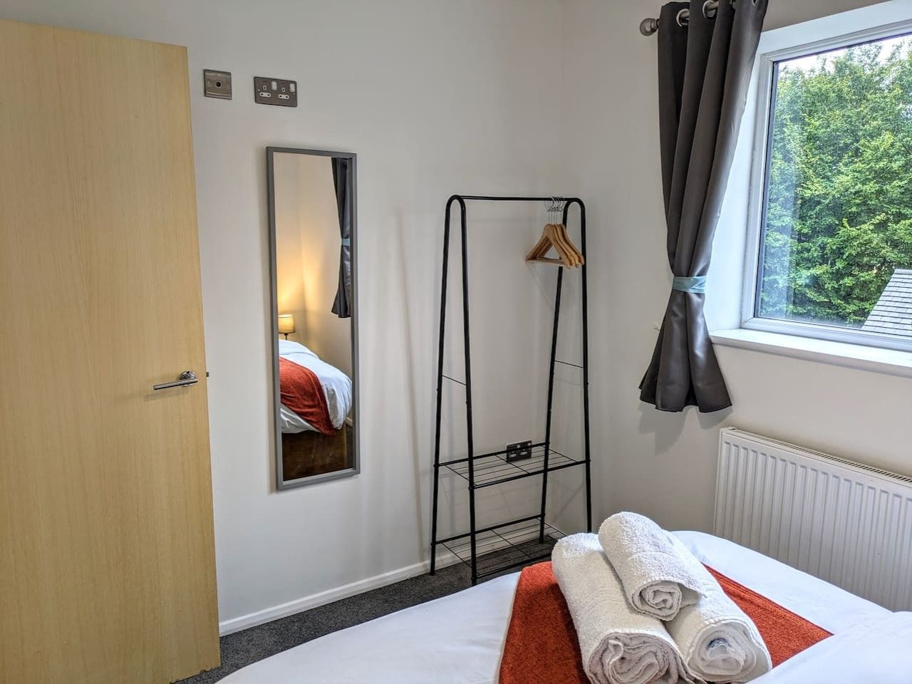 A hotel room with a bed, a mirror, a radiator, a window with curtains, and a towel rack.
