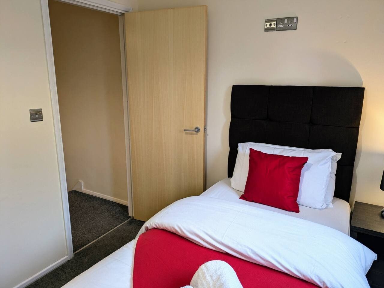 A hotel room with a bed, a bedside table, a nightstand, a door, and a wall socket.