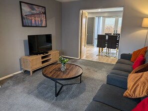 A living room with a TV, a round coffee table, a sofa, and a view into the dining area. - Tempting Telford, 4BR Home, 3 Bathrooms, Parking (Telford and Wrekin)