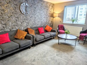 A living room with a grey sofa set, a round coffee table, and a wall clock. - Tempting Telford, 4BR Home, 3 Bathrooms, Parking (Telford and Wrekin)