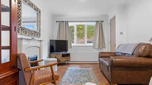A cozy living room with a brown leather sofa, a white armchair, a wooden chair, a TV on a wooden stand, and a large mirror. - Beautiful Renovated Central Home in York with Free Parking | Pass The Keys (York)