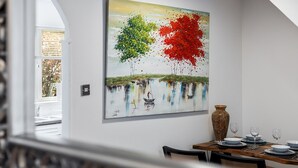 A dining area with a table set for four, a vase, and a painting of trees and water. - Beautiful Renovated Central Home in York with Free Parking | Pass The Keys (York)