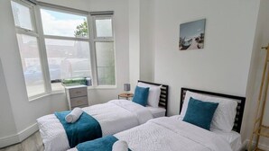 A hotel room with two beds, a window with blinds, a small table with a lamp, and a picture on the wall. - Contractors 4BR Home, Newly Refurb, Nr Centre (West Midlands)