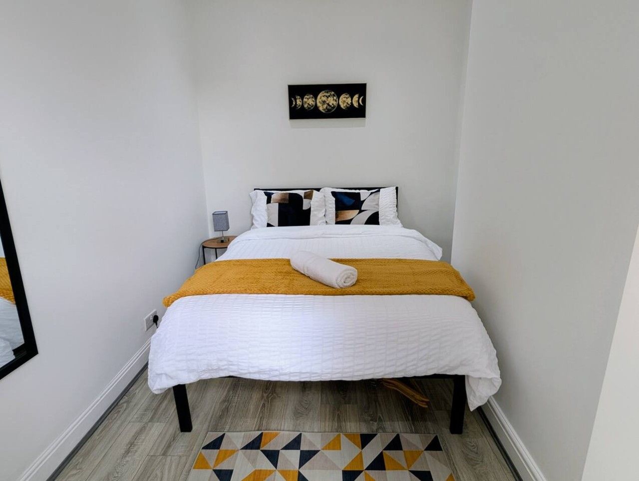 Un lit bien fait, recouvert d’une couverture blanche et jaune, une petite table de chevet avec une lampe, et une œuvre d’art accrochée au mur représentant les différentes phases de la lune.