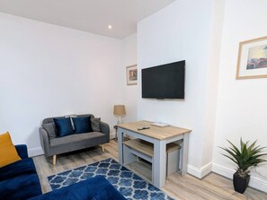 Een moderne woonkamer met een grijze bank, een houten salontafel, een blauw vloerkleed met patroon, een flatscreen televisie aan de muur en een plant in pot.