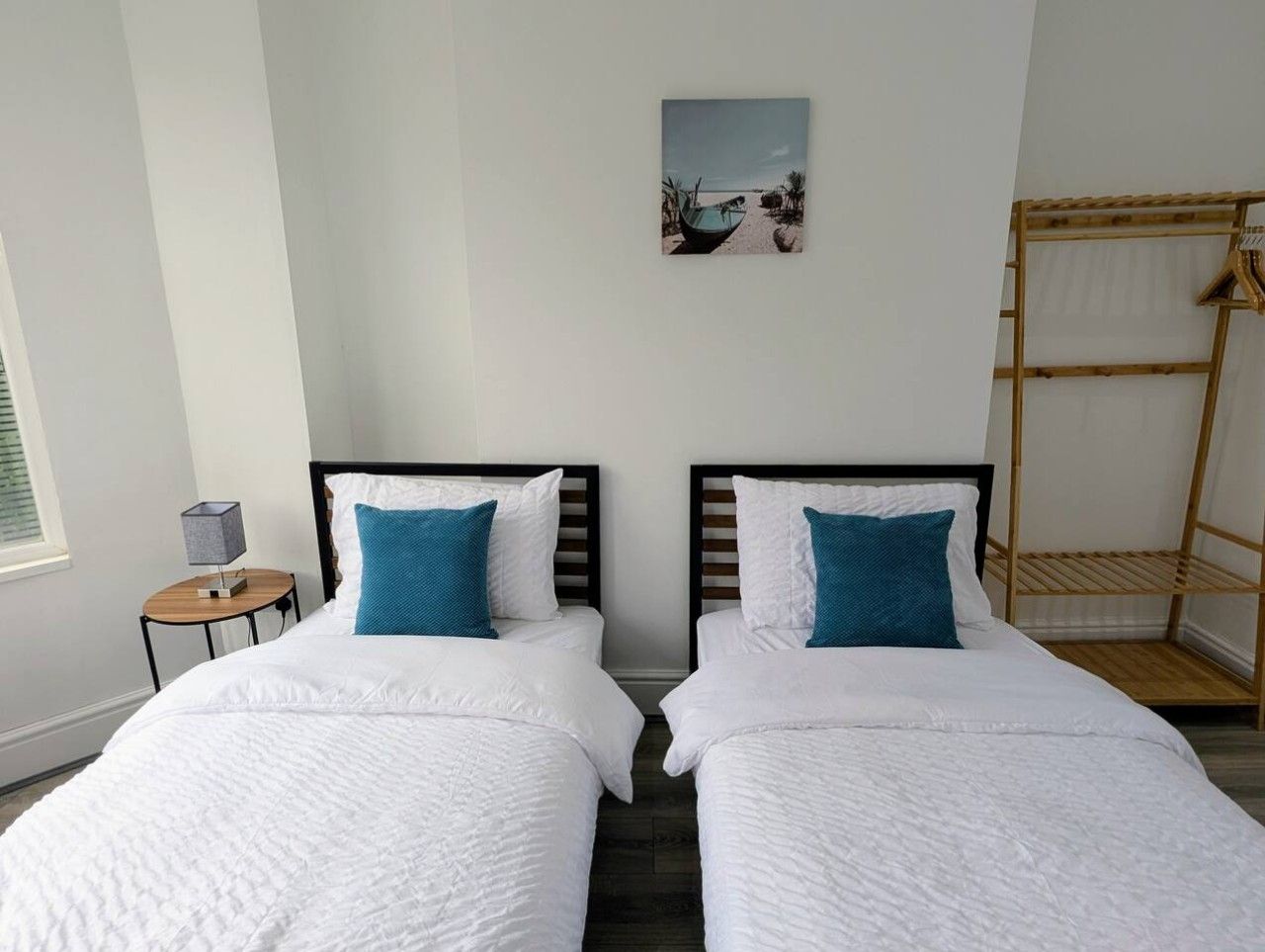 Habitación doble con cama y ropa blanca, dos almohadas azules, una mesita de noche con lámpara y una estantería de madera.