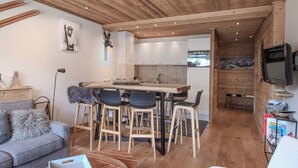Una sala de estar acogedora con techo de madera, una zona de comedor con mesa y sillas de madera, un sofá gris y un televisor a la derecha.