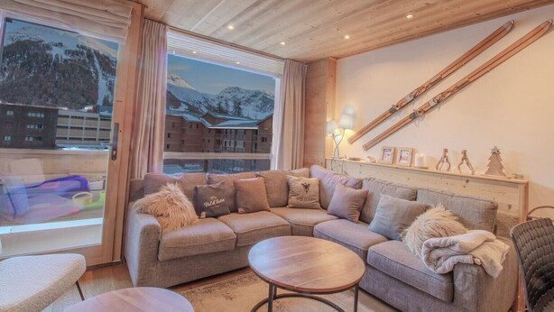 Una sala de estar acogedora con techo de madera, un ventanal con vistas a la montaña, un conjunto de sofás y un perchero para esquíes en la pared.