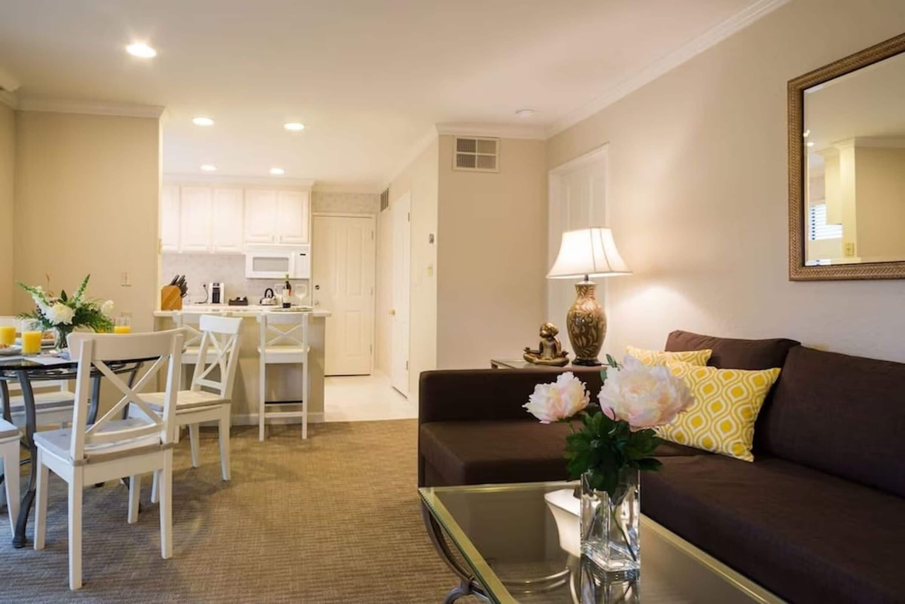 A living room with a brown sofa, a glass coffee table, and a dining area with white chairs and a table.