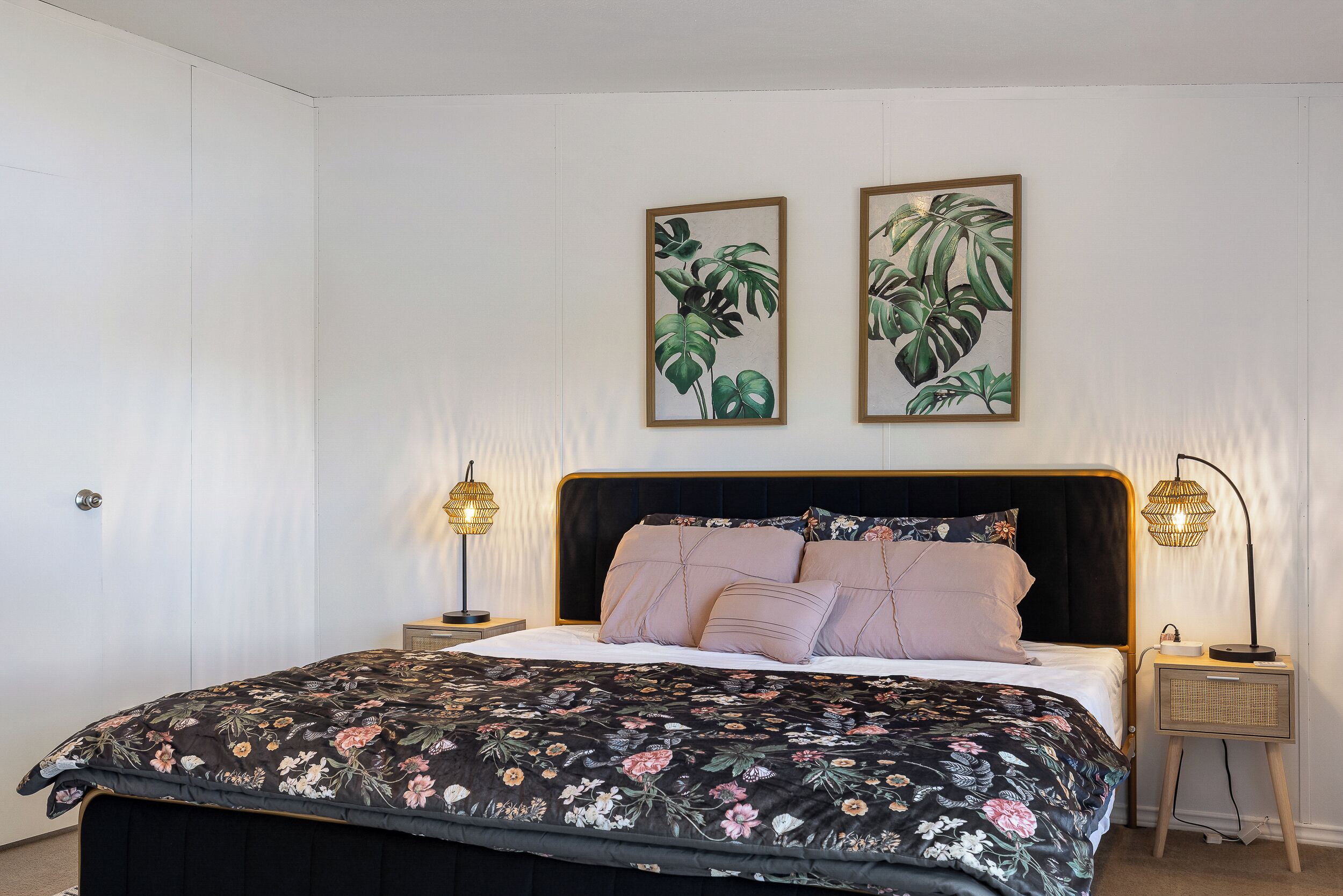 Une chambre à coucher avec un lit noir, une literie à motifs floraux, deux tableaux représentant des plantes accrochés au mur et des lampes de chevet.