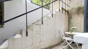 Eine weiße Treppe mit Metallgeländern, ein weißer Tisch mit einer Vase und Früchten sowie ein weißer Stuhl in einem Innenhof mit Backsteinmauer und Fliesenboden.