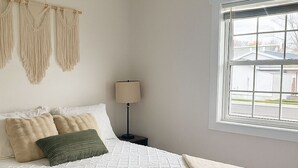 Un dormitorio con cama, lámpara de noche, un tapiz de macramé y una ventana con vistas.