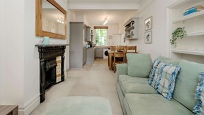 A living room with a fireplace, a sofa, a dining table, and a kitchen in the background. - Modern 2 bed Flat - Hammersmith Grove (London)