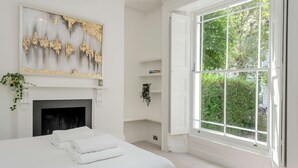 A bedroom with a bed, a fireplace, a large window, and a wall-mounted shelf. - Modern 2 bed Flat - Hammersmith Grove (London)